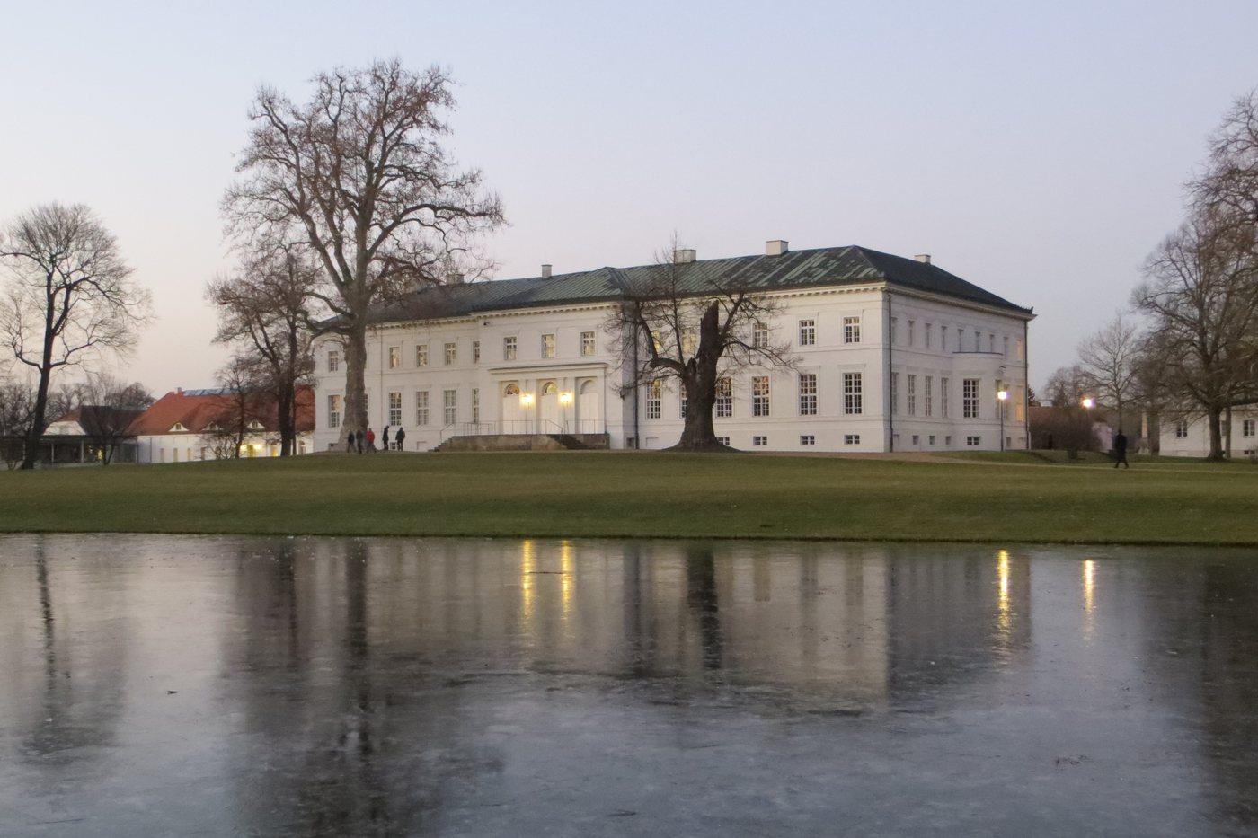 Schloss Neuhardenberg in der Dämmerung mit erleuchtetem Portal, davor der zugefrorene Teich